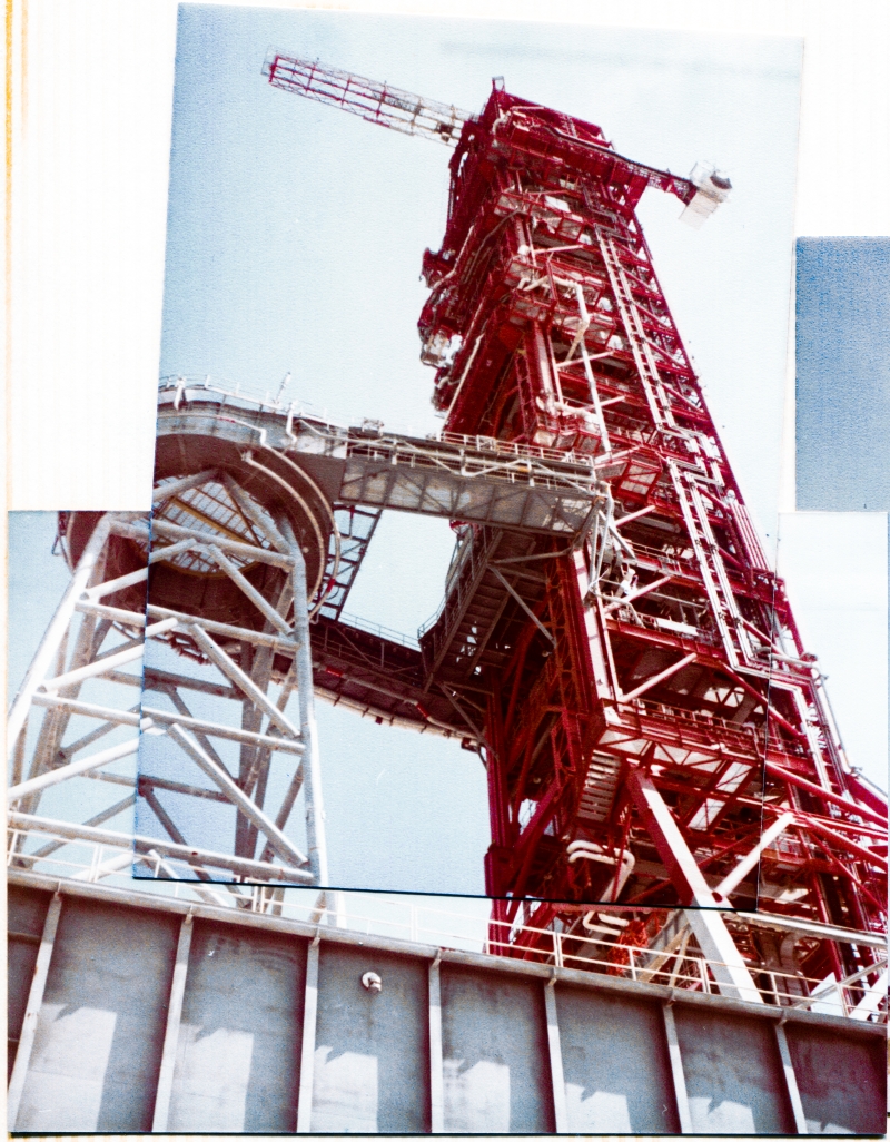 Mobile Launcher 1 looms over 400 feet into the sky above you as you stand on the dirt beneath it at the Park Site north of the VAB. Next to the red Launch Umbilical Tower (LUT) the gray steel structure of the Milk Stool can be seen. The Milk Stool is what the Saturn 1B’s which flew from this ML sat on top of, permitting the use of the ML for a much smaller launch vehicle than the Saturn V’s which it was originally designed and constructed for pre-launch servicing. Photograph by James MacLaren.